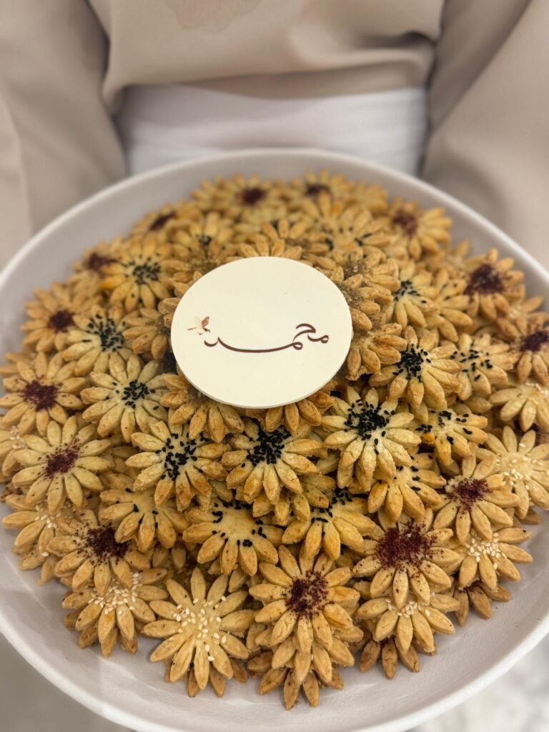 An elegant platter of Arabic petit fours by Hala Cookies, beautifully shaped like flowers and topped with sesame seeds, featuring a custom disc with elegant Arabic calligraphy, perfect for Eid and cultural celebrations in Doha.