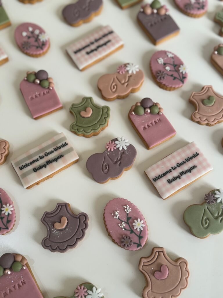 An assortment of custom baby shower cookies by Hala Cookies, featuring "Welcome to Our World Baby Mayan" messages and elegant floral and onesie designs in muted colors, perfect for welcoming a new baby in Doha.