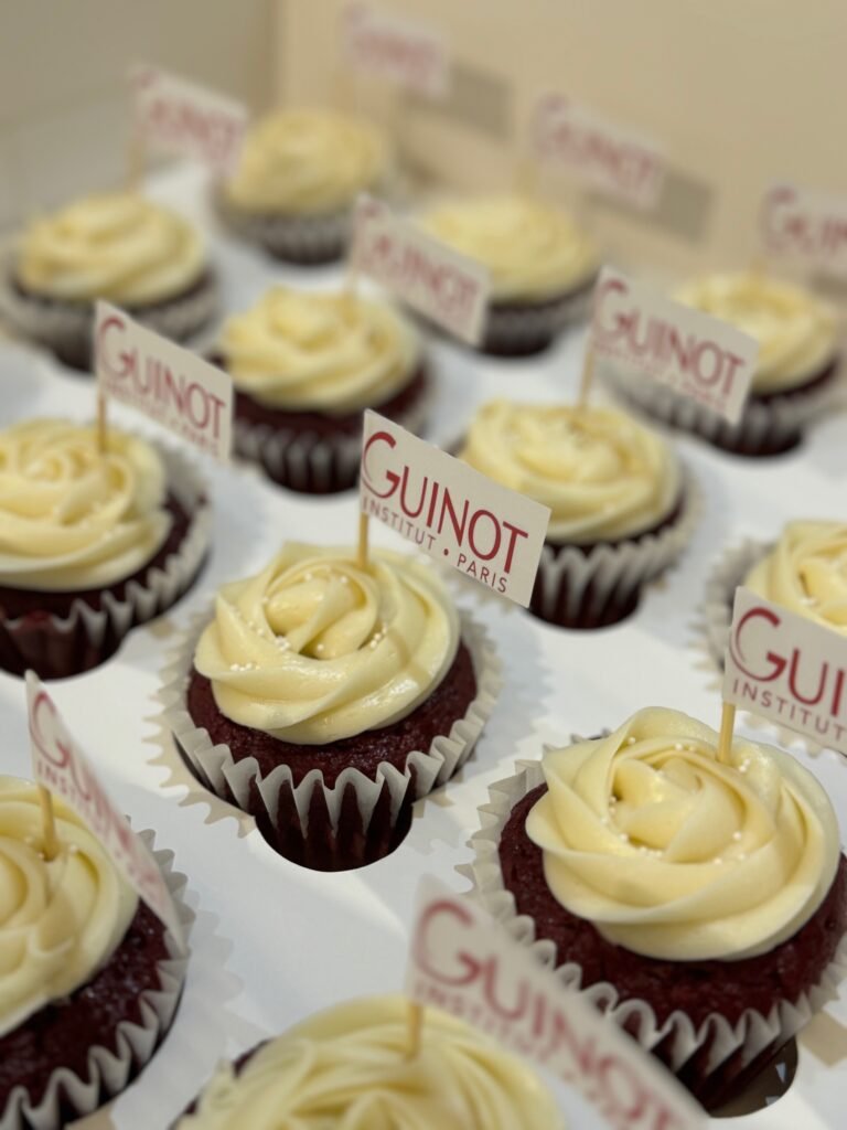 Custom red velvet corporate cupcakes by Hala Cookies, featuring individual "Guinot Institut Paris" edible flags, ideal for business events and client gifts in Doha.