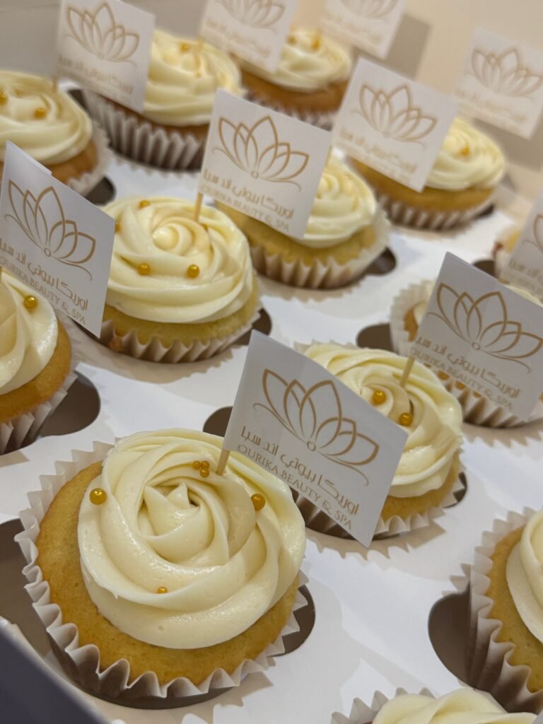 A box of custom corporate cupcakes by Hala Cookies, featuring white frosting rosettes, gold sprinkles, and branded flags for Ourika Beauty & Spa, perfect for corporate events and client gifts in Doha.