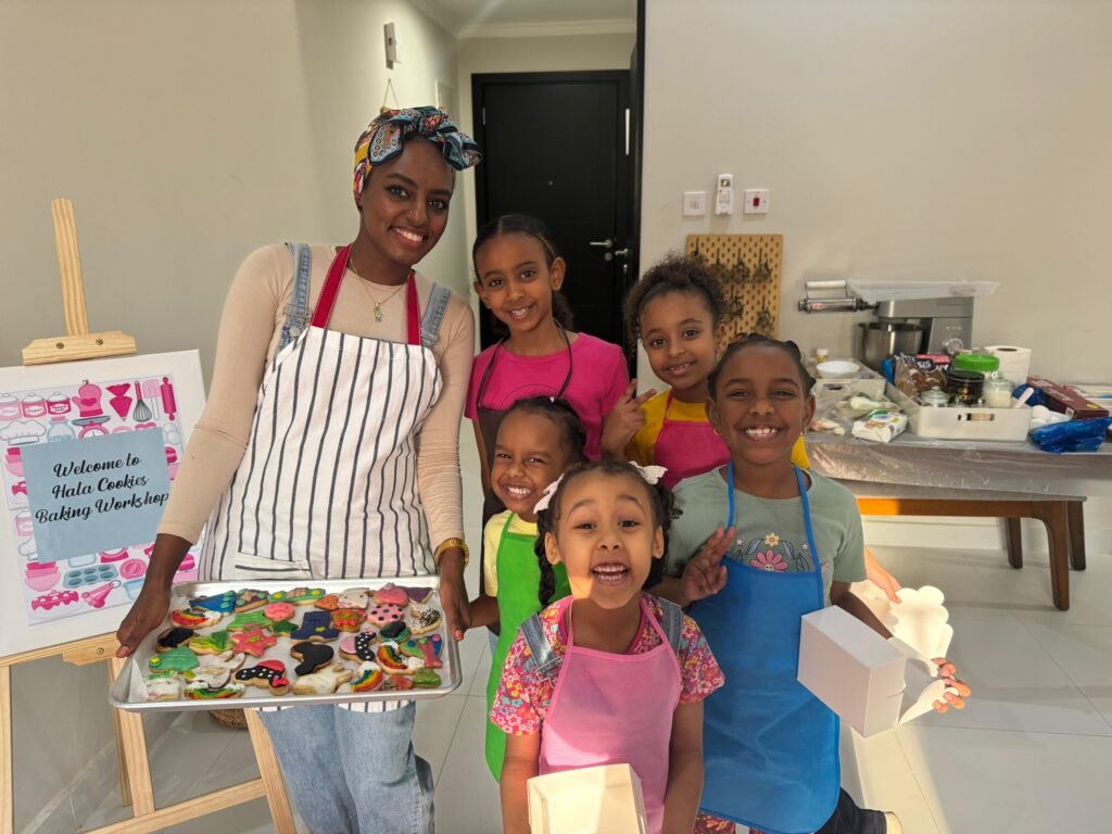 Hala, the founder of Hala Cookies, smiling with a group of happy children at a kids' baking workshop in Doha, holding a tray of colorful, freshly decorated cookies.